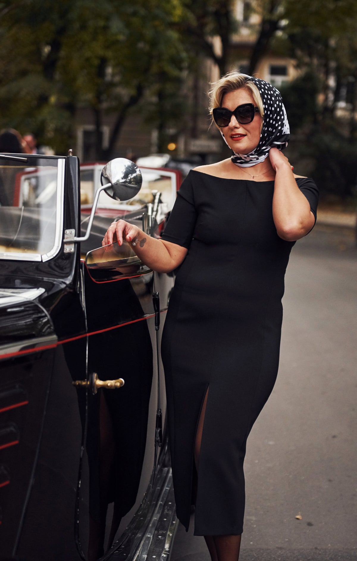 Woman in a black dress standing next to an old-fashioned car with trees in the background